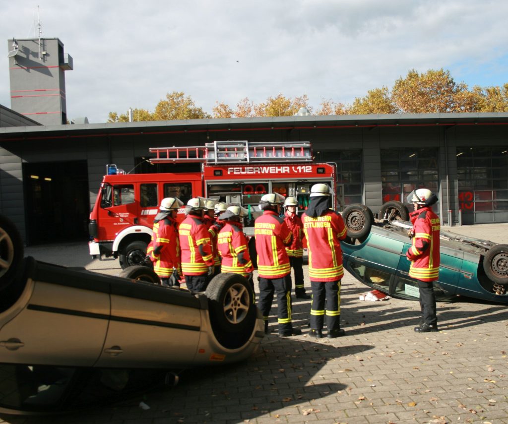Zwei PKW auf Seite liegend, eine Gruppe Feuerwehrleute mit Ausbilder daneben stehend 